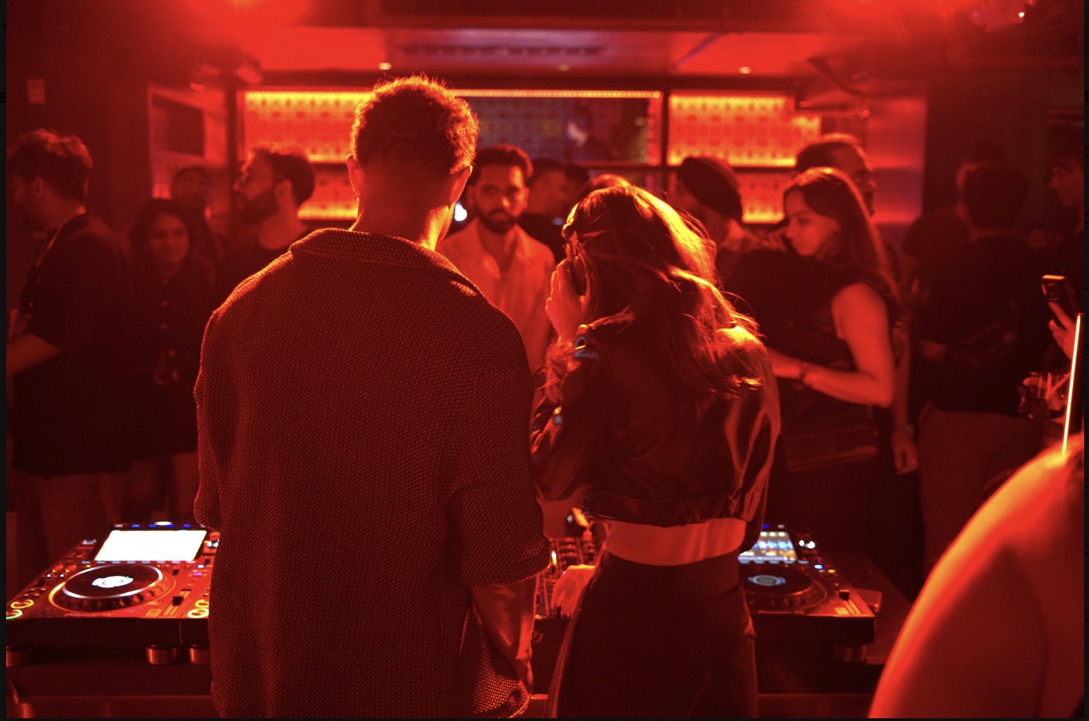 Two DJs at the decks facing the crowd under red light at 16 Sola